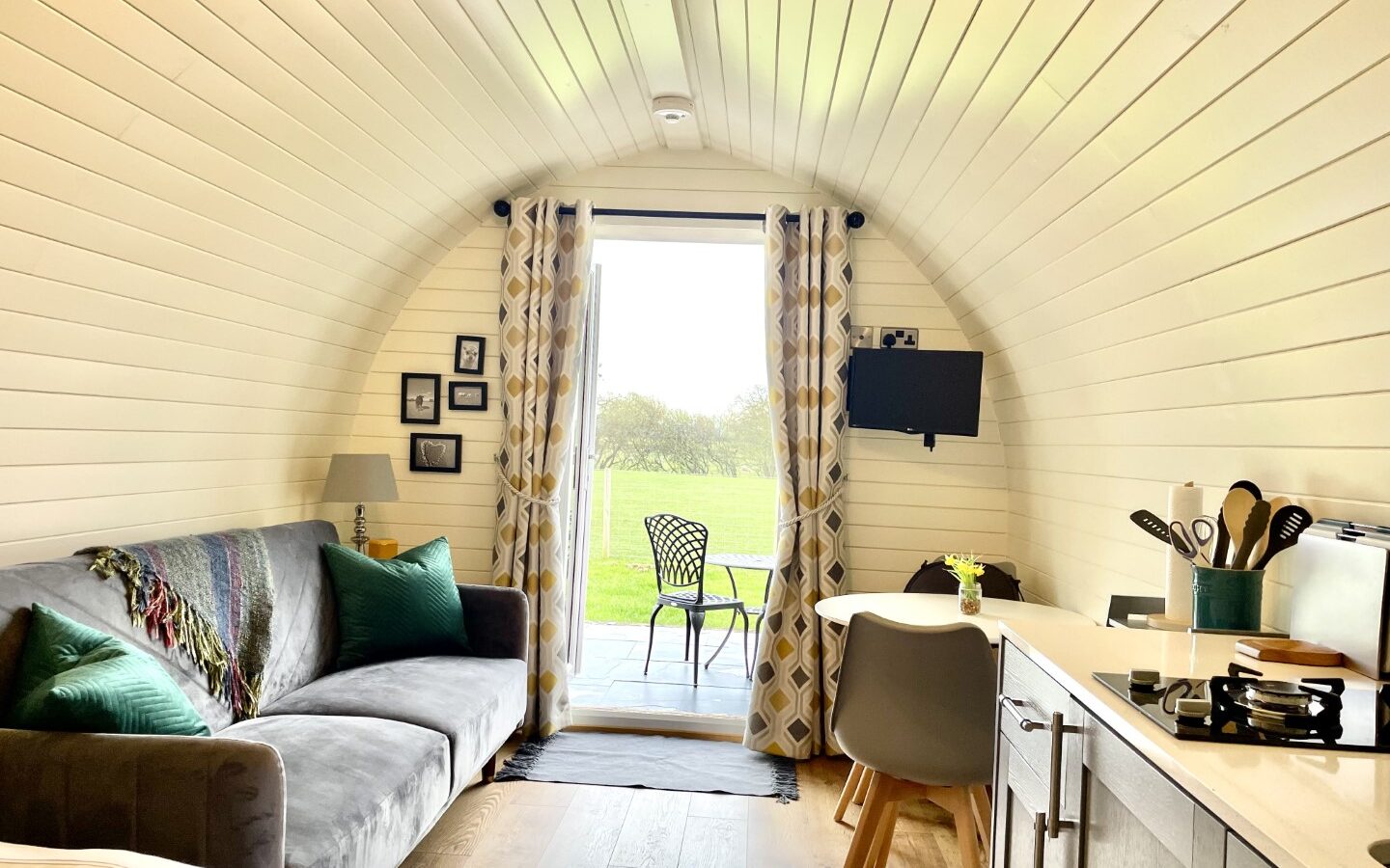 A cozy, modern tiny home at Abbey Farm with white, wooden walls and an arched ceiling. The space features a sofa, small dining table, TV, and kitchen area. Large glass doors with patterned curtains open to a patio with a chair, perfect for glamping and overlooking a lush, green outdoor view.