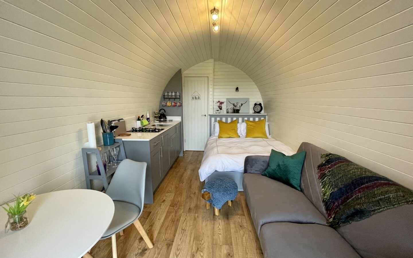 A cozy, modern tiny home at Abbey Farm with a wooden interior features a kitchenette with gray cabinets, a small dining table with two chairs, a bed with white and yellow bedding, and a gray sofa adorned with colorful cushions. This inviting glamping retreat is well-lit and welcoming.