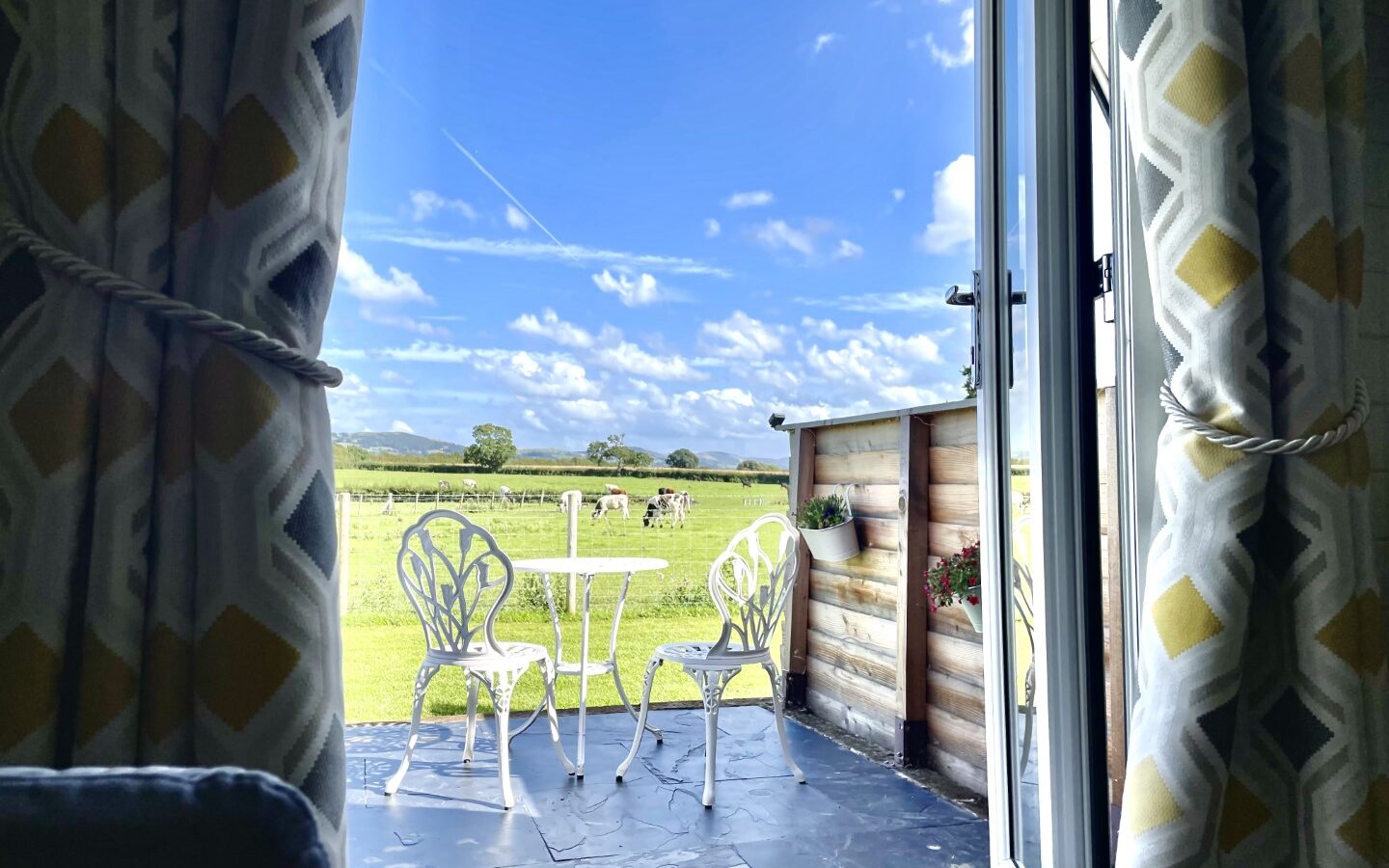 A set of double doors open to reveal a cozy patio with a small white table and two matching chairs, perfect for glamping at Abbey Farm, overlooking green fields and a bright blue sky with scattered clouds. Patterned curtains are tied back, framing the serene outdoor scene.