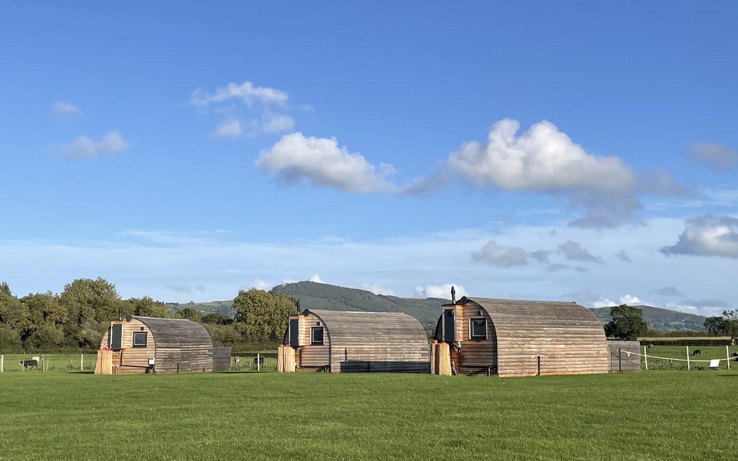 Three small wooden cabins with curved roofs are situated on the lush green field of Abbey Farm under a bright blue sky with sparse clouds. Behind the cabins, there are trees and hills in the distance, perfect for a serene glamping experience.