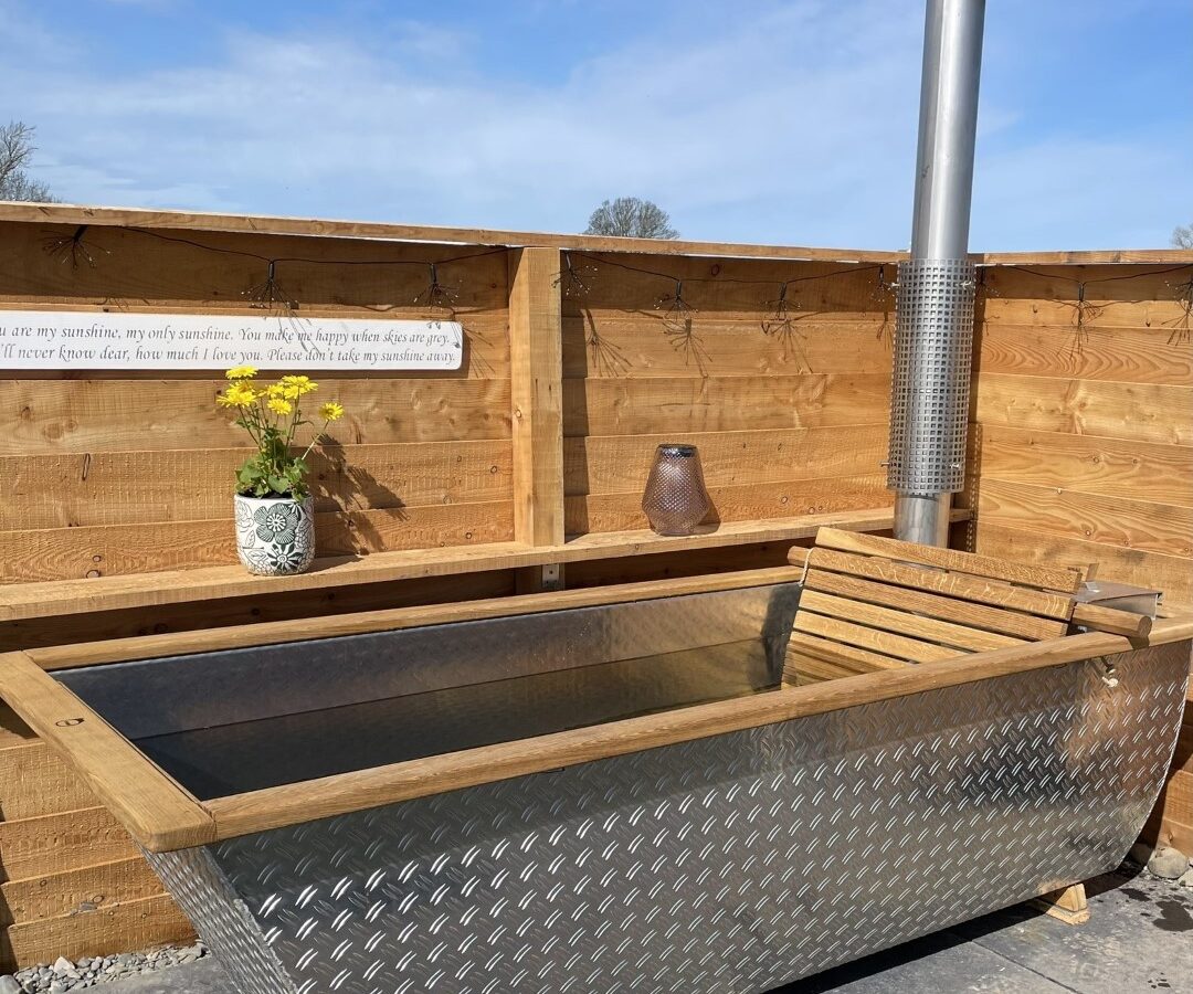 A metal outdoor hot tub with wooden trim and a tall chimney is set against a wooden fence on a stone-tiled patio at Abbey Farm. A vase with yellow flowers and a lantern are placed on the fence, and a framed sign is attached to the upper part of the fence, perfect for glamping on a sunny day.