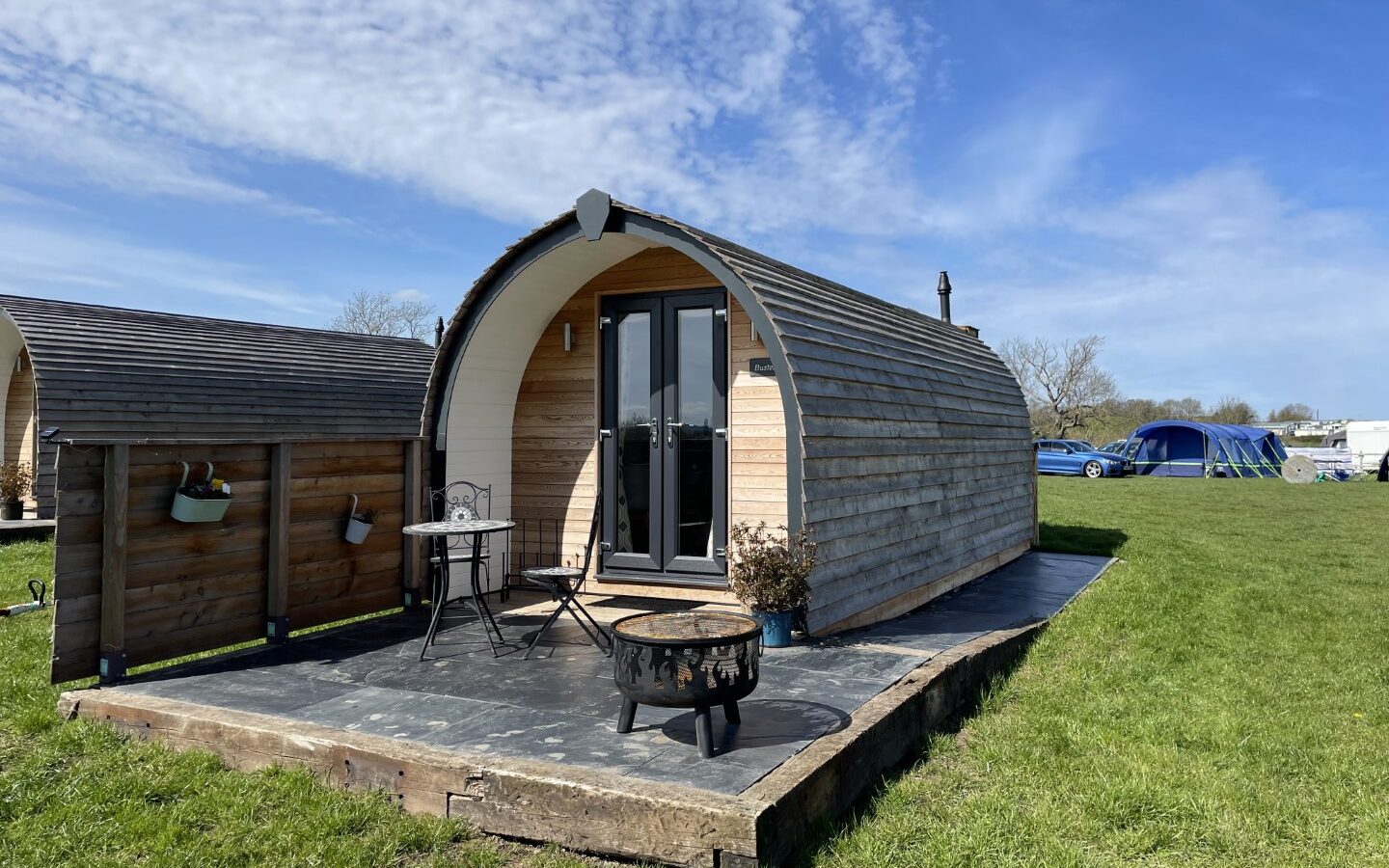 A wooden pod cabin with a curved roof sits on a grassy area under a blue sky. It features a small patio with a table, two chairs, and a fire pit. Nearby at Abbey Farm Glamping, a blue tent is set up, and a few cars are parked in the background.