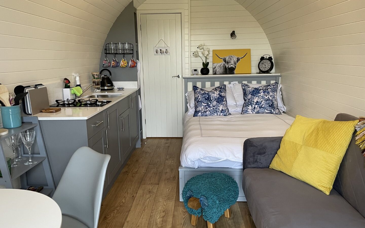 A cozy, arched tiny home perfect for glamping at Abbey Farm features a bed with blue and white floral pillows, a gray sofa with yellow cushions, a small kitchenette with a stove and kettle, and a dining set with gray chairs. The wooden floor contrasts with the white paneled walls and a cow painting decorates the space.