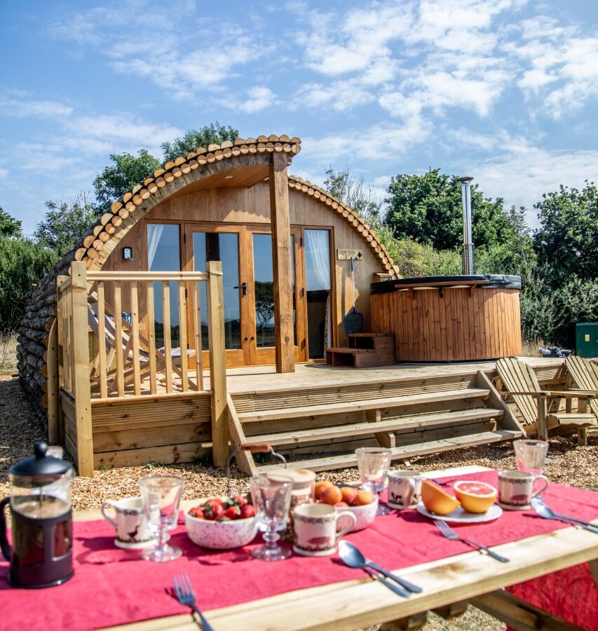 A wooden cabin with a Barnhorn-inspired curved roof sits in a scenic outdoor setting. In front of the cabin is a small deck with steps leading up to it and a round hot tub, perfect for glamping. A picnic table in the foreground is set with breakfast items, including coffee, fruit, and pastries. The sky is clear with some clouds.
