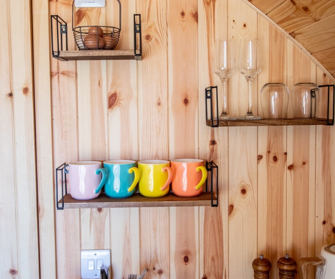 A cozy wooden kitchen at Barnhorn Glamping features a curved ceiling, black metal shelves holding two wine glasses, three jars, and a basket of onions. Colorful mugs (yellow, pink, blue, green) adorn the middle shelf. A cutlery holder with utensils sits on the counter alongside salt and pepper mills.