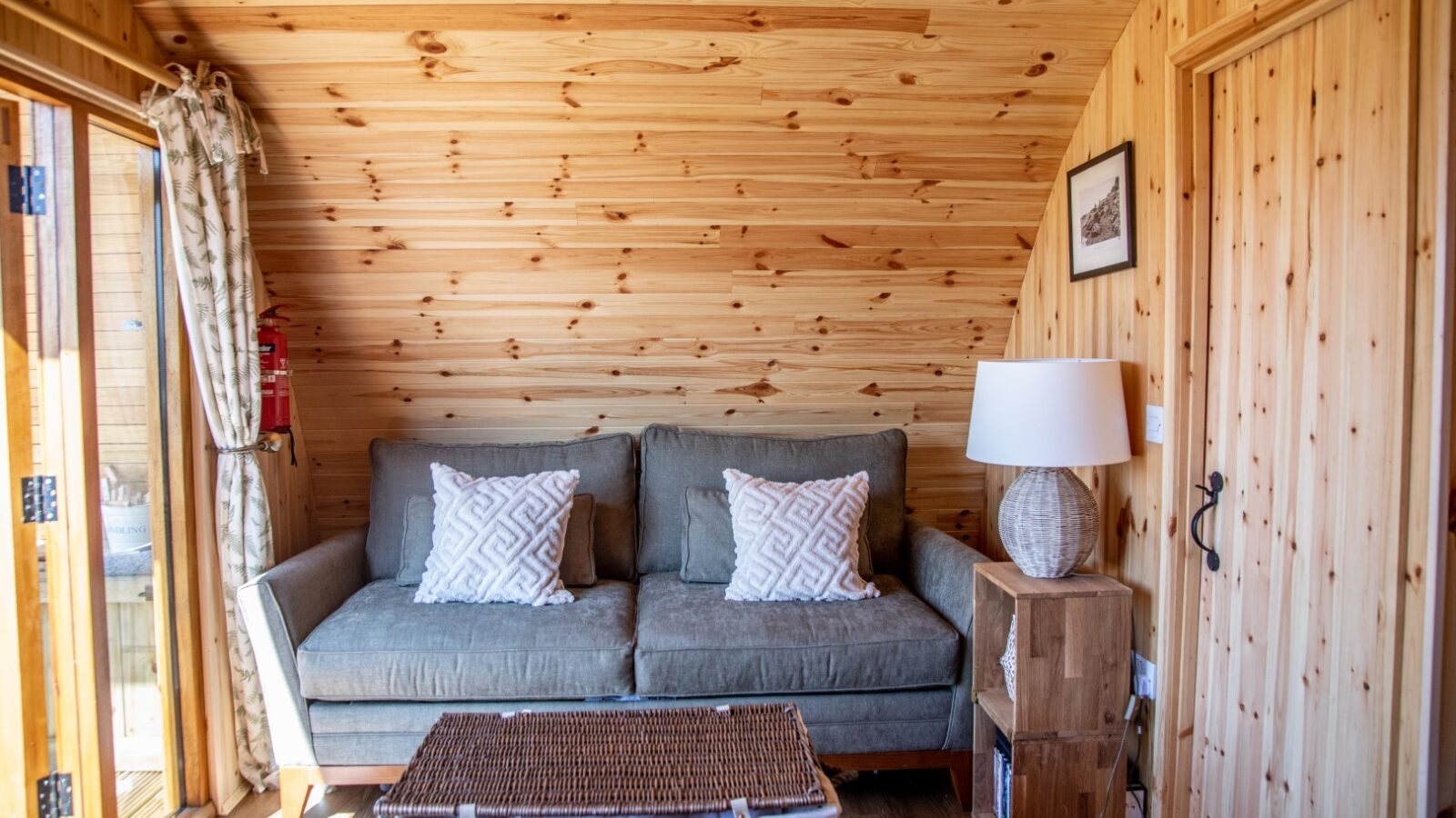 A cozy wooden cabin interior with a rustic feel perfect for glamping. The space features a gray cushioned sofa adorned with patterned white pillows. A wicker coffee table sits in front, and a wooden side table with a white lamp is next to the sofa. The walls and ceiling are made of pine wood.