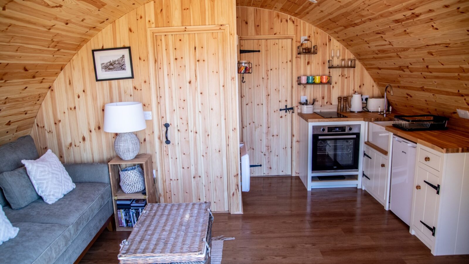 A cozy, rustic cabin interior with wooden walls and ceiling perfect for glamping. It features a small kitchen area with a stove, oven, refrigerator, and shelves holding colorful mugs. There is a sofa with cushions, a side table with a lamp, and a wicker chest in the center. A framed picture hangs on the wall.