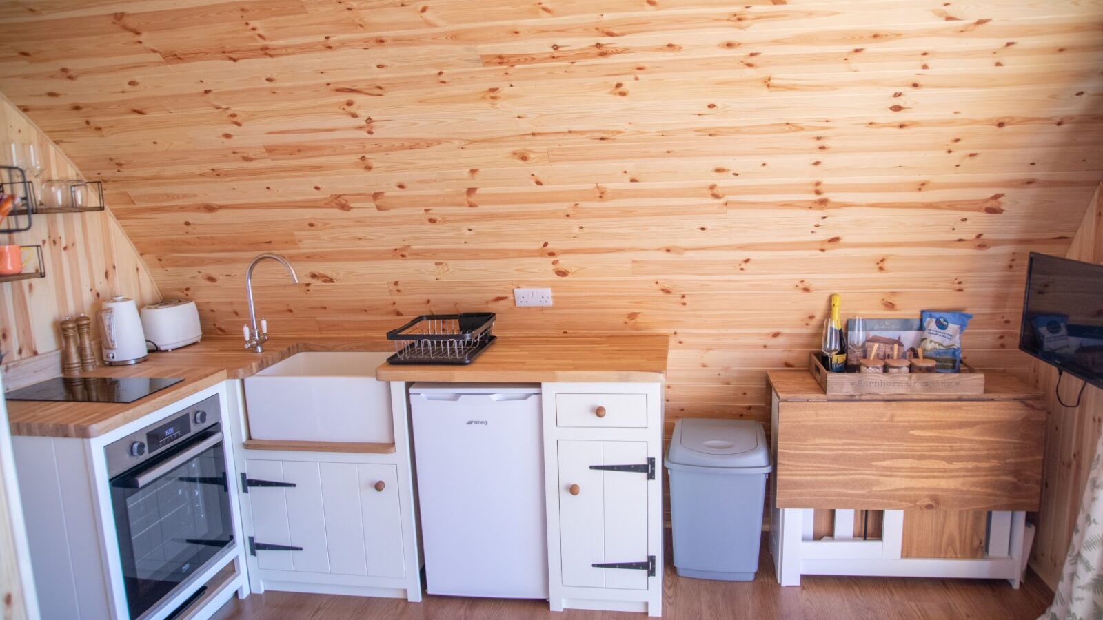 A cozy wooden kitchen with shiny wooden walls and floors, perfect for a glamping getaway. It features a small white oven, a sink with a drying rack, and a mini fridge. A countertop holds a wine bottle, glasses, and snacks. A small trash bin sits next to the counter. Adjacent is a wooden table with a TV on it.