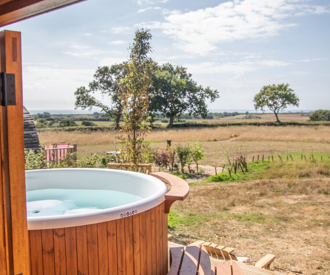 A wooden hot tub with curved steps is on a deck just outside a Barnhorn cabin door, overlooking a grassy field with scattered trees under the clear blue sky. The open door frame provides a view from inside the cabin out to the serene glamping landscape.