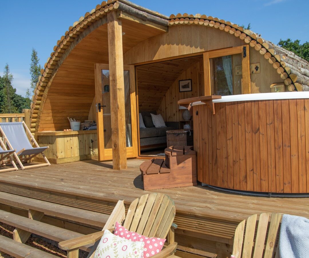 A cozy wooden cabin with a curved roof and outdoor deck, perfect for glamping, featuring two Adirondack chairs with patterned cushions. The cabin has large glass doors, a hot tub next to the entrance, and a pair of deck chairs to the left. The clear blue sky completes the Barnhorn escape.