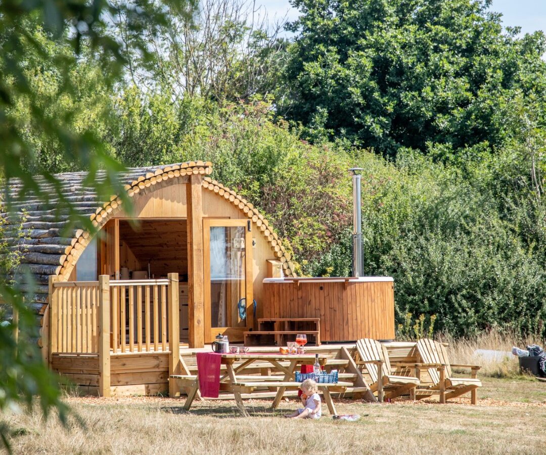 A cozy, wooden pod-style cabin at Barnhorn Glamping sits in a grassy field with surrounding trees and clear blue sky. In front, there's a picnic table, outdoor hot tub, and a child sitting on a wooden bench. Bushes and greenery provide a natural, serene backdrop for the ultimate glamping experience.