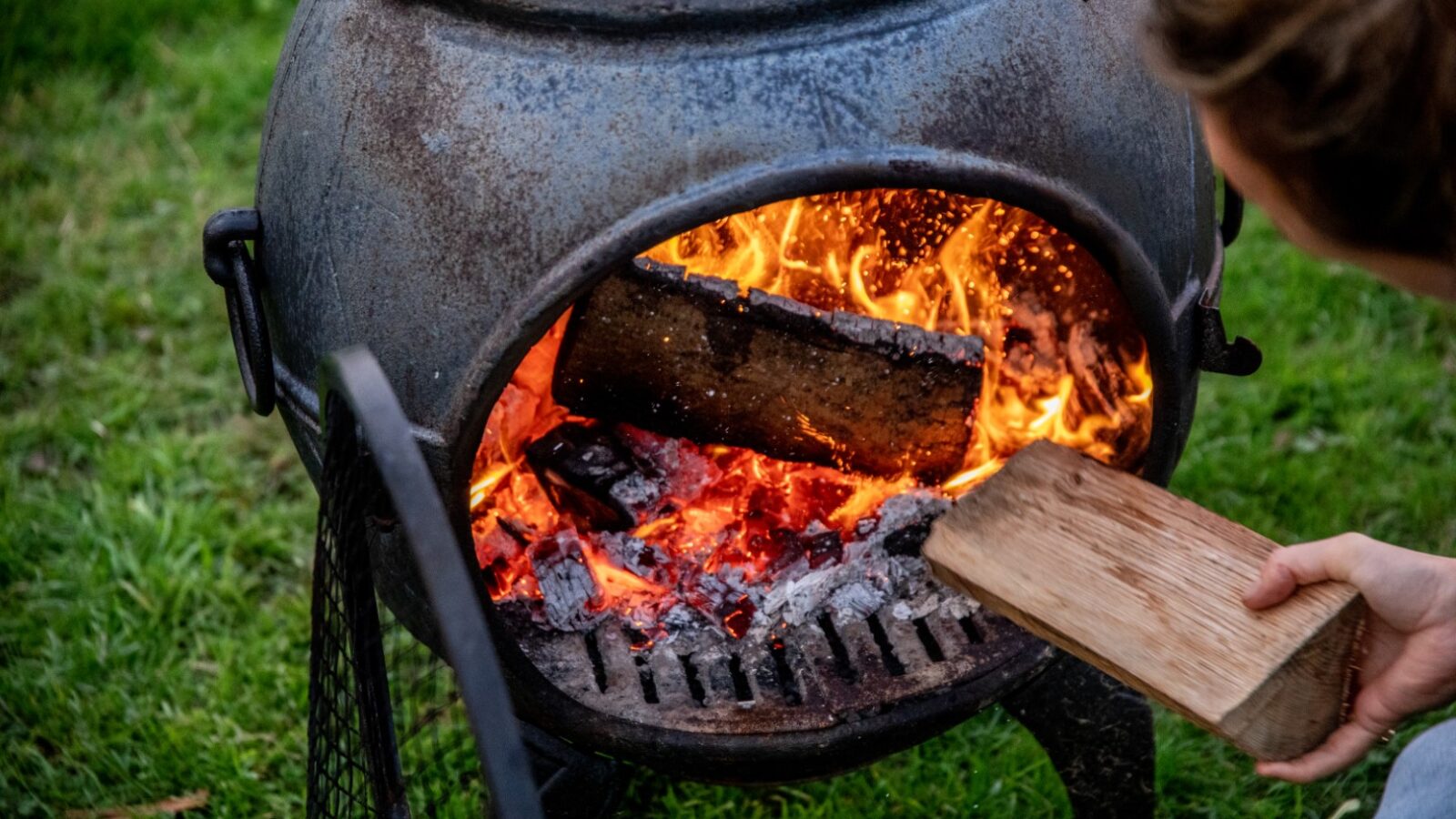 A person is placing a wooden log onto a fire inside a metal fire pit, enjoying the glamping experience. The fire pit stands on a grassy area, with flames and embers clearly visible. The person's hand is holding the log, while the rest of their body is partially out of the frame.