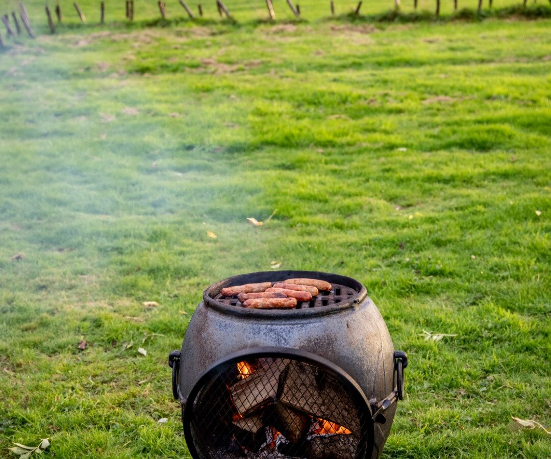 Small outdoor barbecue grill with steaks cooking on top, set on a grassy field with trees and a fence in the background. Smoke rises from the grill, blending into the serene landscape. The sky is overcast, giving this glamping scene a peaceful, countryside ambiance perfect for relaxation.