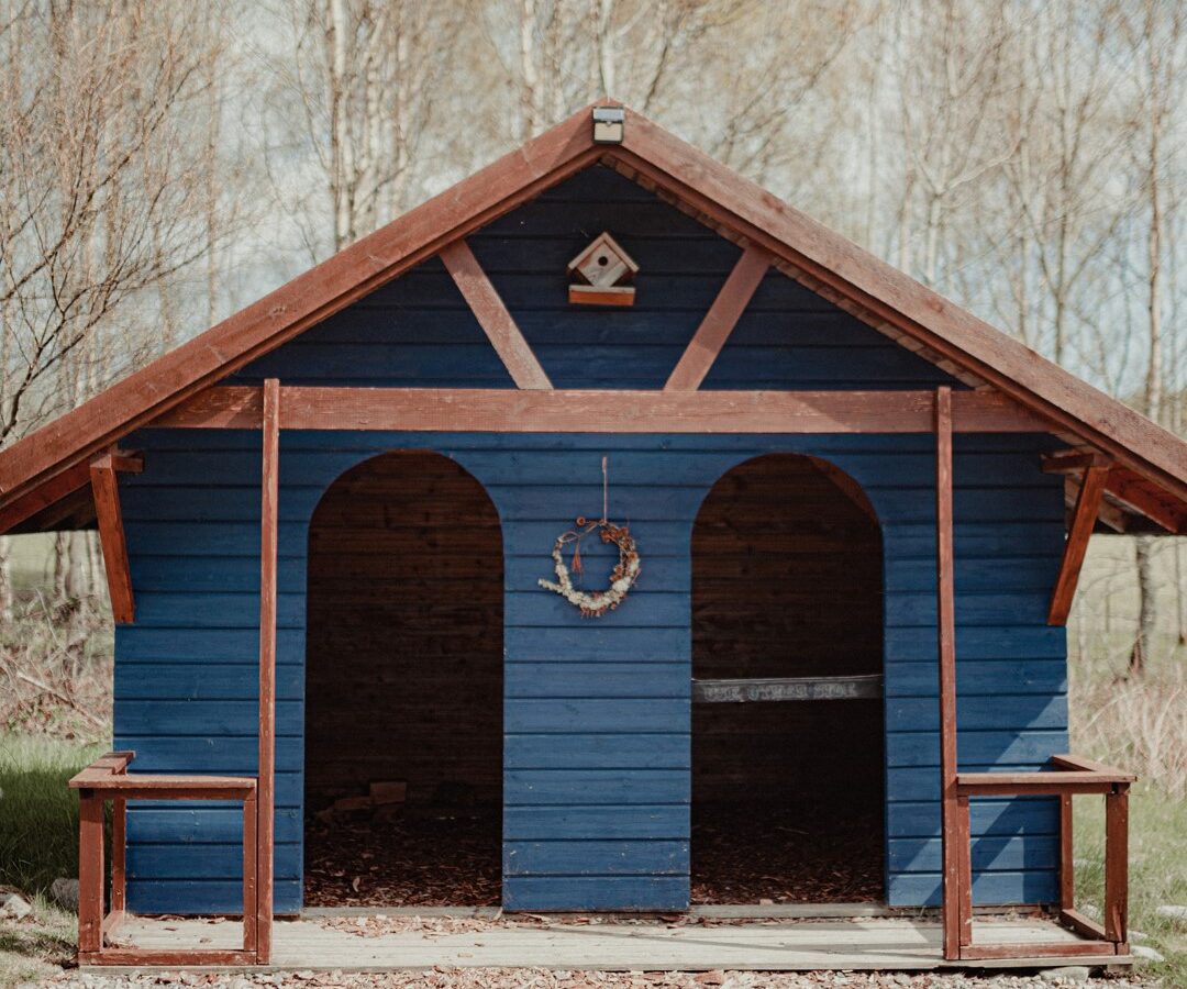 A blue wooden cabin with a gabled roof, adorned with a small birdhouse at the top and a wreath hanging between two archways at the front. This charming retreat by Big Sky Lodges is set in a wooded area with leafless trees in the background.