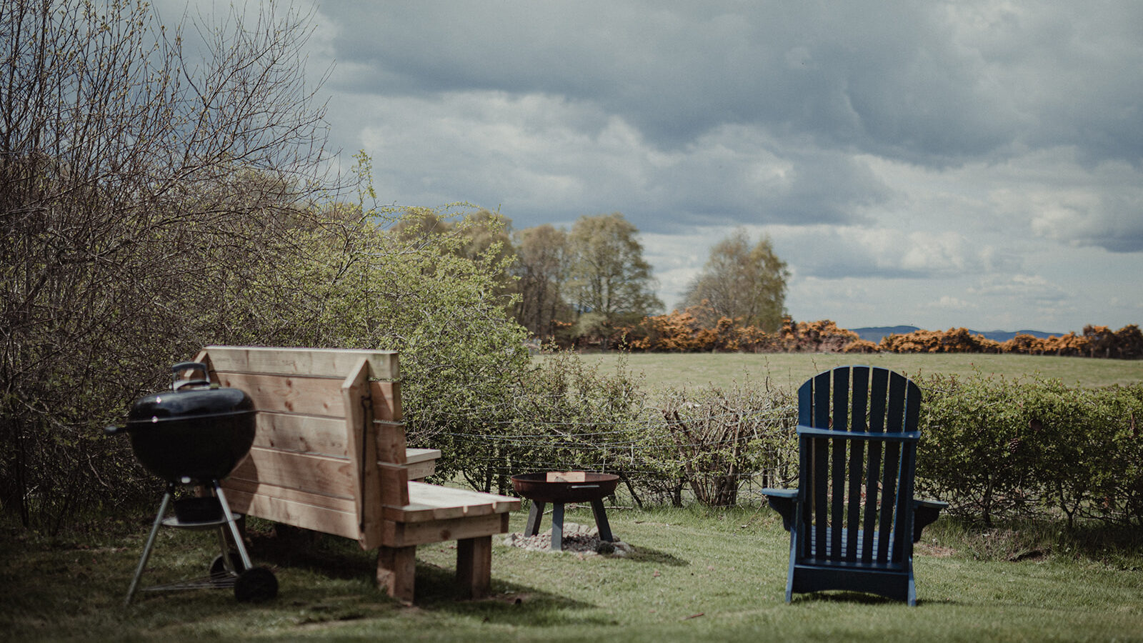 A serene backyard scene at Big Sky Lodges features a wooden bench, a blue Adirondack chair, and a black charcoal grill. The area is surrounded by lush greenery under a cloudy sky, with trees and yellow flowering bushes in the background.