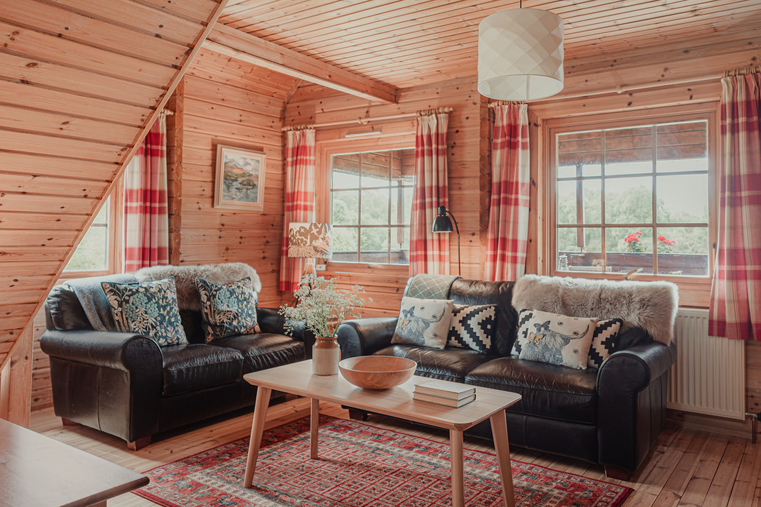 A cozy wooden living room, reminiscent of rustic lodges, features two dark leather sofas adorned with patterned cushions and faux fur throws. A light wood coffee table holds a bowl and a book, while red and white checkered curtains frame the windows. Above, a lamp hangs from the ceiling, highlighting the Big Sky decor.