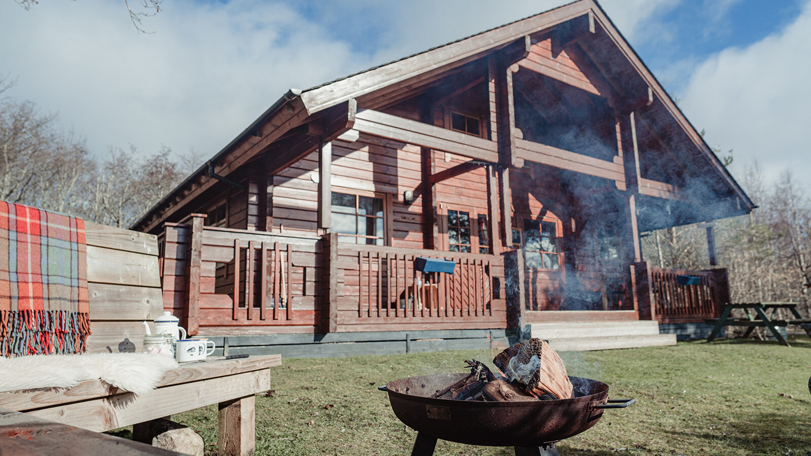 A wooden cabin with a large porch sits in a scenic outdoor setting at Big Sky Lodges. In the foreground, there's a fire pit with burning logs, a wooden bench adorned with a red plaid blanket, and a small white coffee maker. The sky is partly cloudy.