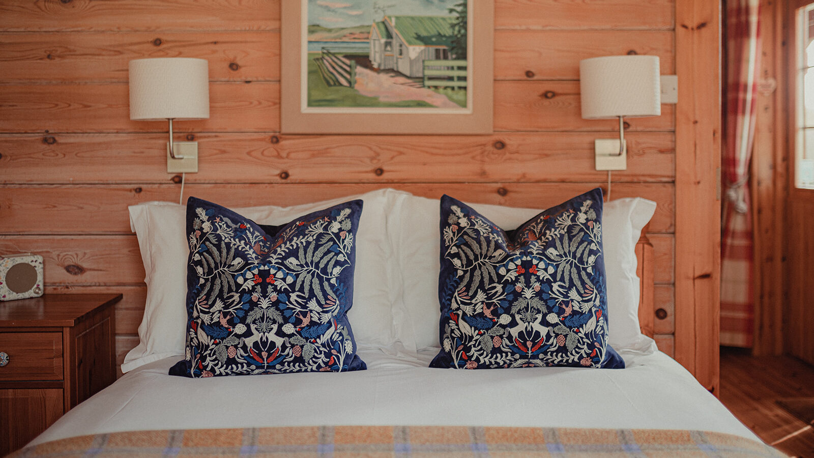 A cozy bedroom in Big Sky with a wooden interior features a neatly made bed with white linens and two decorative pillows with intricate blue and red patterns. Above the bed, a framed painting of a landscape hangs on the wall, flanked by two wall-mounted lamps, evoking the warmth of rustic lodges.