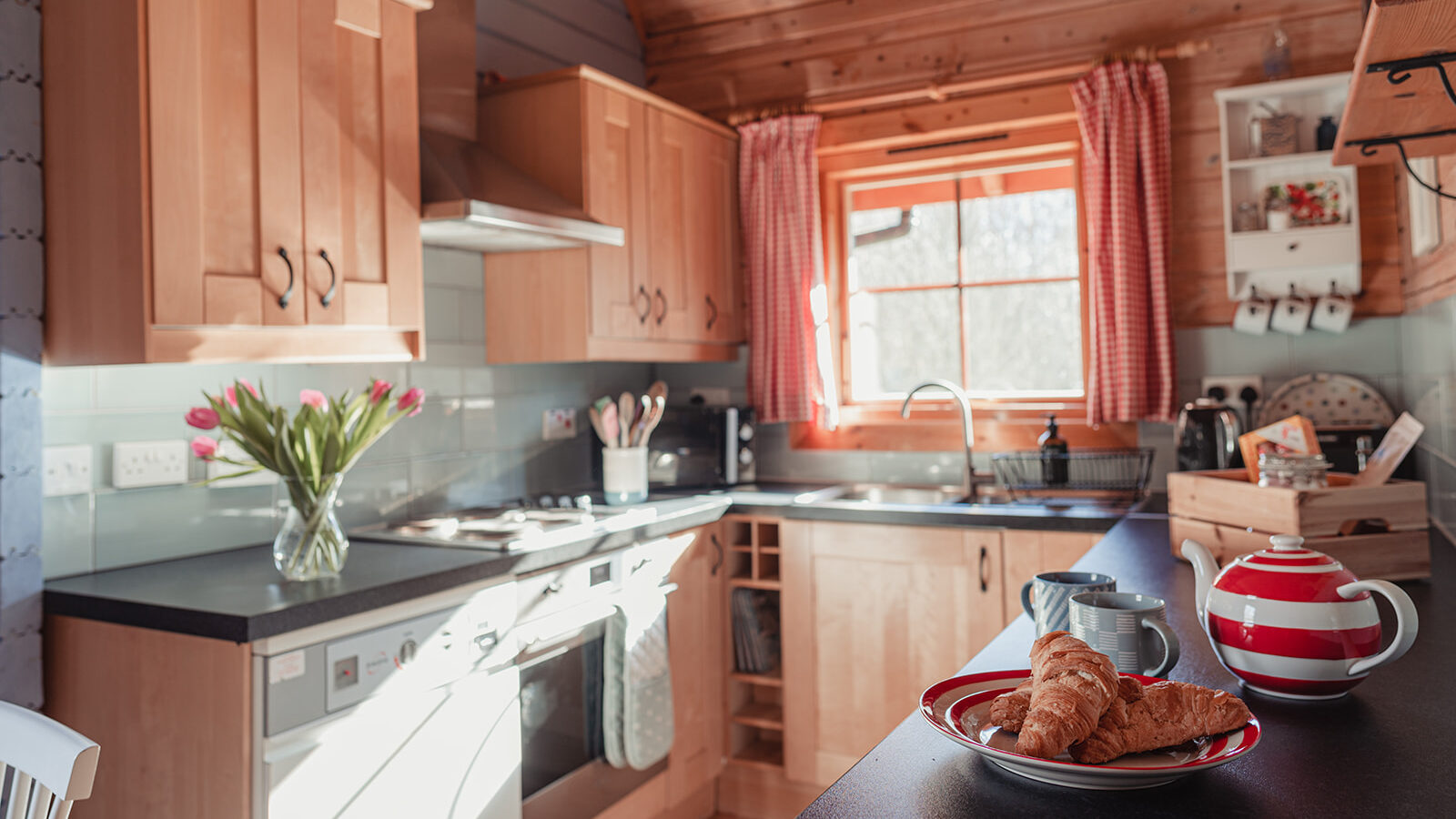 A cozy kitchen with wooden cabinets and a black countertop reminiscent of Big Sky lodges. On the counter, there are croissants on a plate, a striped teapot, and a cup. A vase of tulips sits near a window with red-checked curtains. Various kitchen items are visible, giving a homey feel.