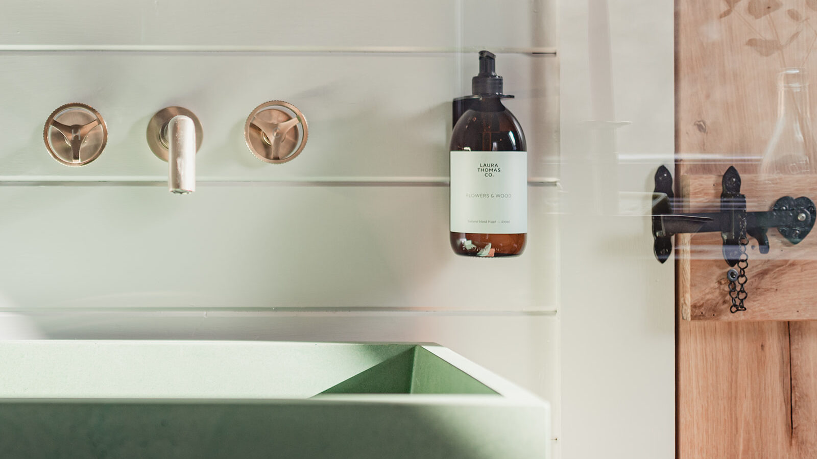 A contemporary bathroom sink area at Big Sky Lodges features a green rectangular basin, mounted under a metallic faucet set against light-colored wooden panels. A brown bottle of liquid soap hangs beside the faucet. In the background, a glass partition with a lit candle can be seen.