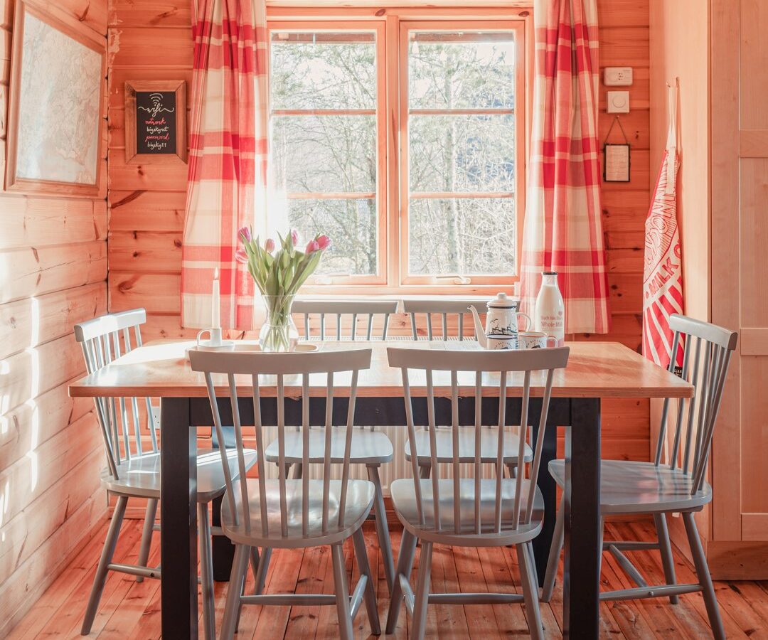 Cozy wooden dining area at Big Sky Lodges with a rectangular table and six light gray chairs. The table holds a vase of tulips and a few items. Red and white plaid curtains frame a window bringing in natural light, complementing the warm wood tones of the walls and floor.