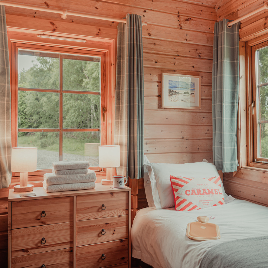 A cozy wooden cabin bedroom at Big Sky Lodges features two windows covered by gray curtains. A single bed with white sheets and a striped accent pillow sits on the right, next to a wooden dresser with stacked towels and lit lamps. Outside, lush greenery is visible.