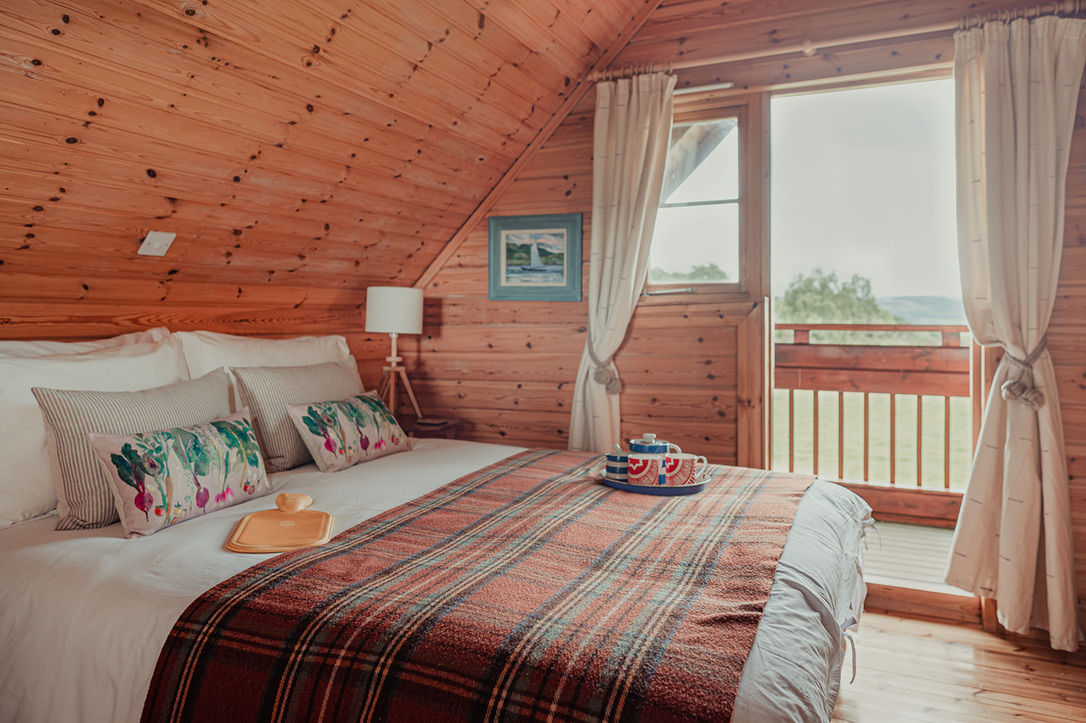 A cozy attic bedroom with wooden walls and ceiling is available at Big Sky Lodges. It features a neatly made double bed with colorful pillows and a plaid blanket. A tray with tea and a teapot sits on the bed. The room has a window with curtains leading to a small balcony with a scenic view.
