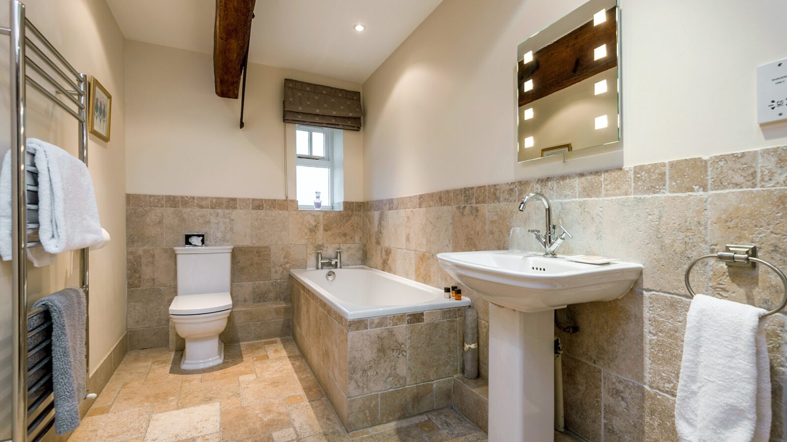 A modern bathroom at Broadgate Farm Cottages boasts beige tiled floors and half-tiled walls. It includes a white toilet, a bathtub with a towel rack nearby, and a pedestal sink with a mirror above. The room features recessed lighting, a wooden beam on the ceiling, and a small window with a Roman shade curtain above the bathtub.