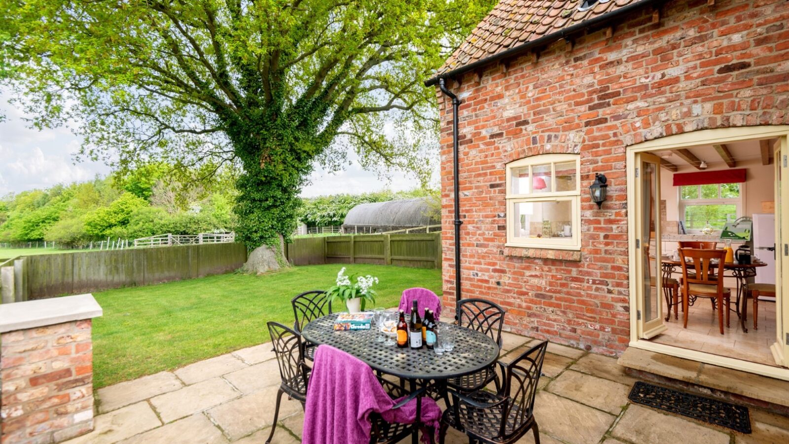 A charming Broadgate Farm cottage with an outdoor patio featuring a round table set with drinks and snacks. A purple blanket drapes over a chair. The patio overlooks a lush, green lawn with a large tree and a fence in the background. The cottage door is open, showing an inviting interior.