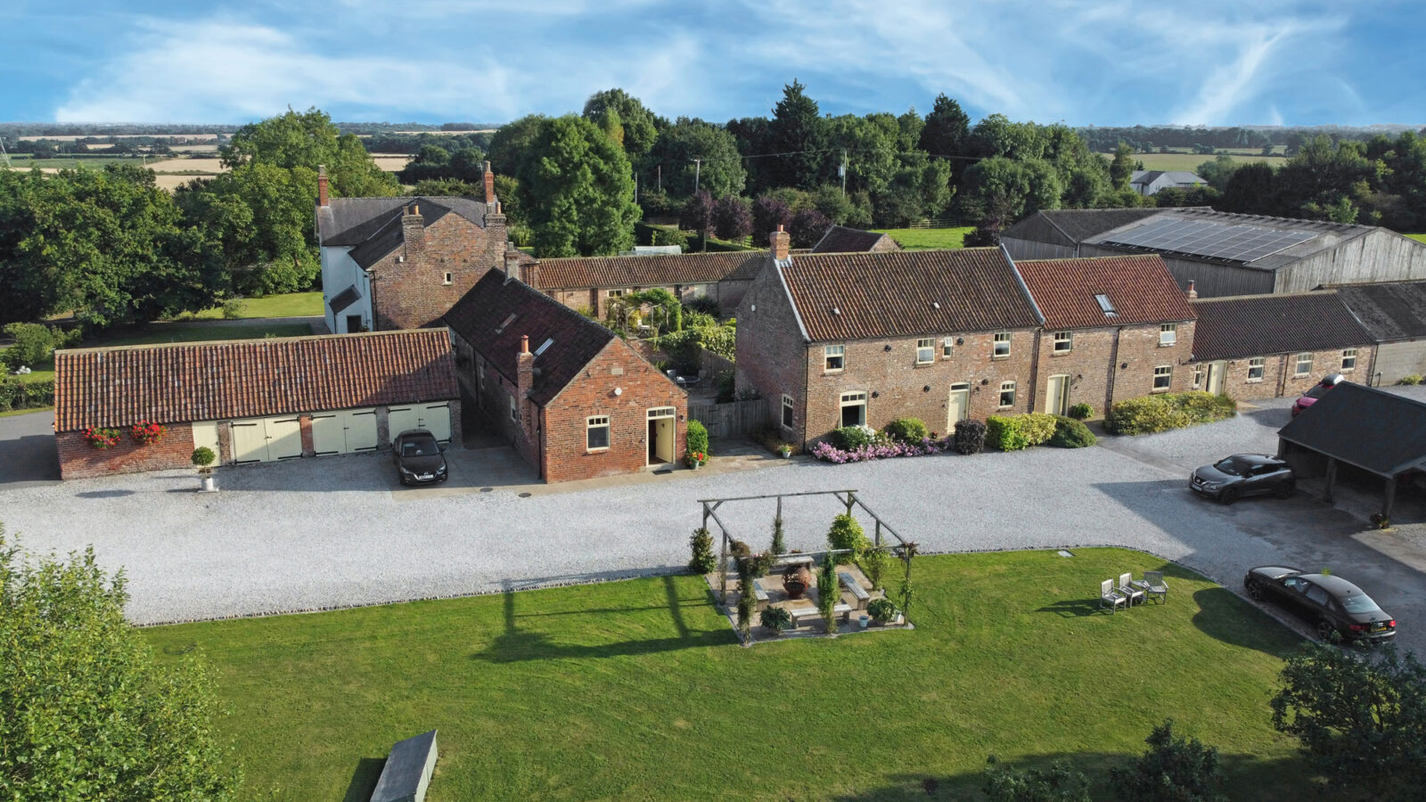 Aerial view of Broadgate Farm Cottages, a rustic property featuring multiple red-brick buildings with tiled roofs, surrounded by greenery. The central yard is covered in gravel, with parked cars and a garden area containing a pergola. Farmland extends into the distance.