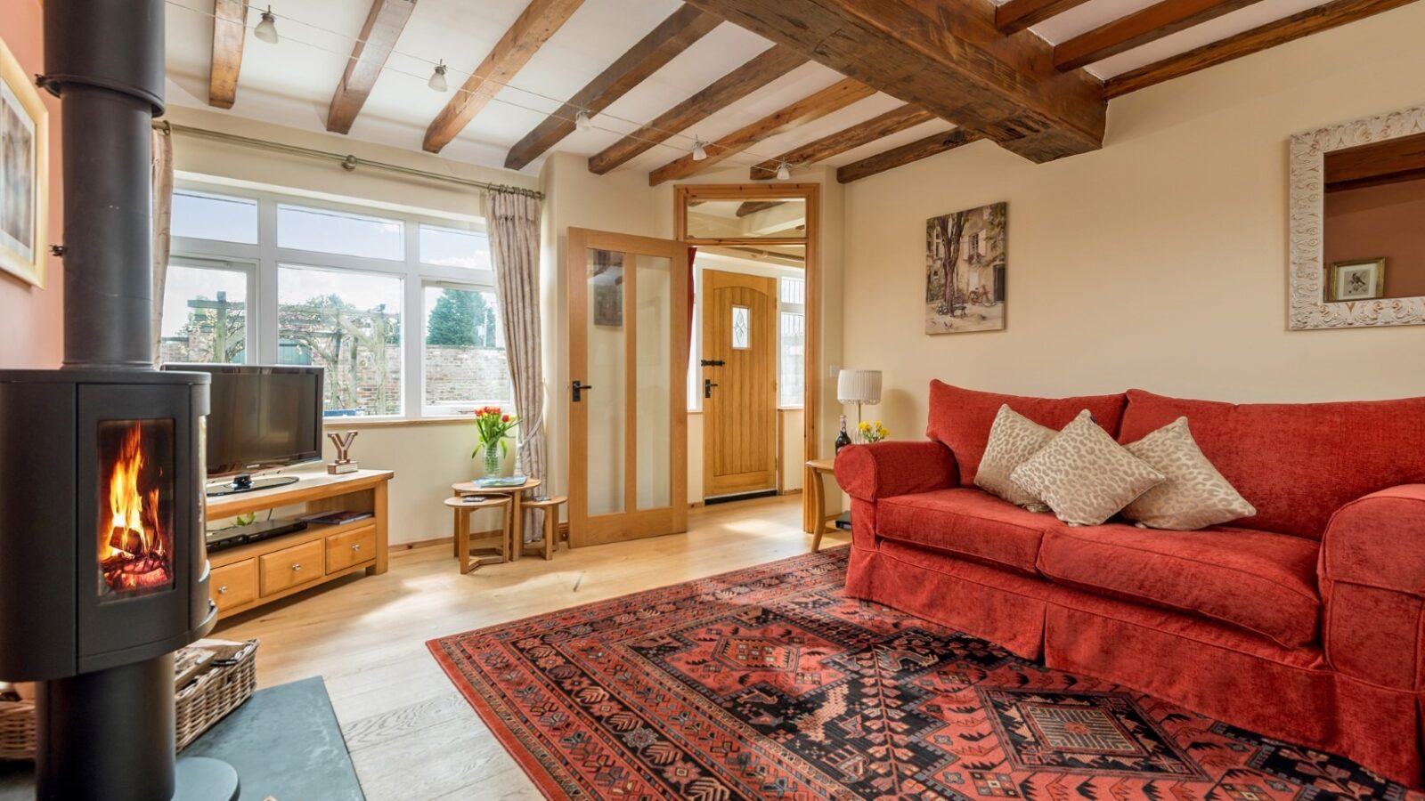 A cozy living room at Broadgate Farm Cottages with a wooden-beamed ceiling, a red sofa adorned with cushions, and a large patterned red rug on the floor. A lit fireplace to the left, a TV on a wooden stand, and a window allowing natural light to fill the space complete this charming retreat.