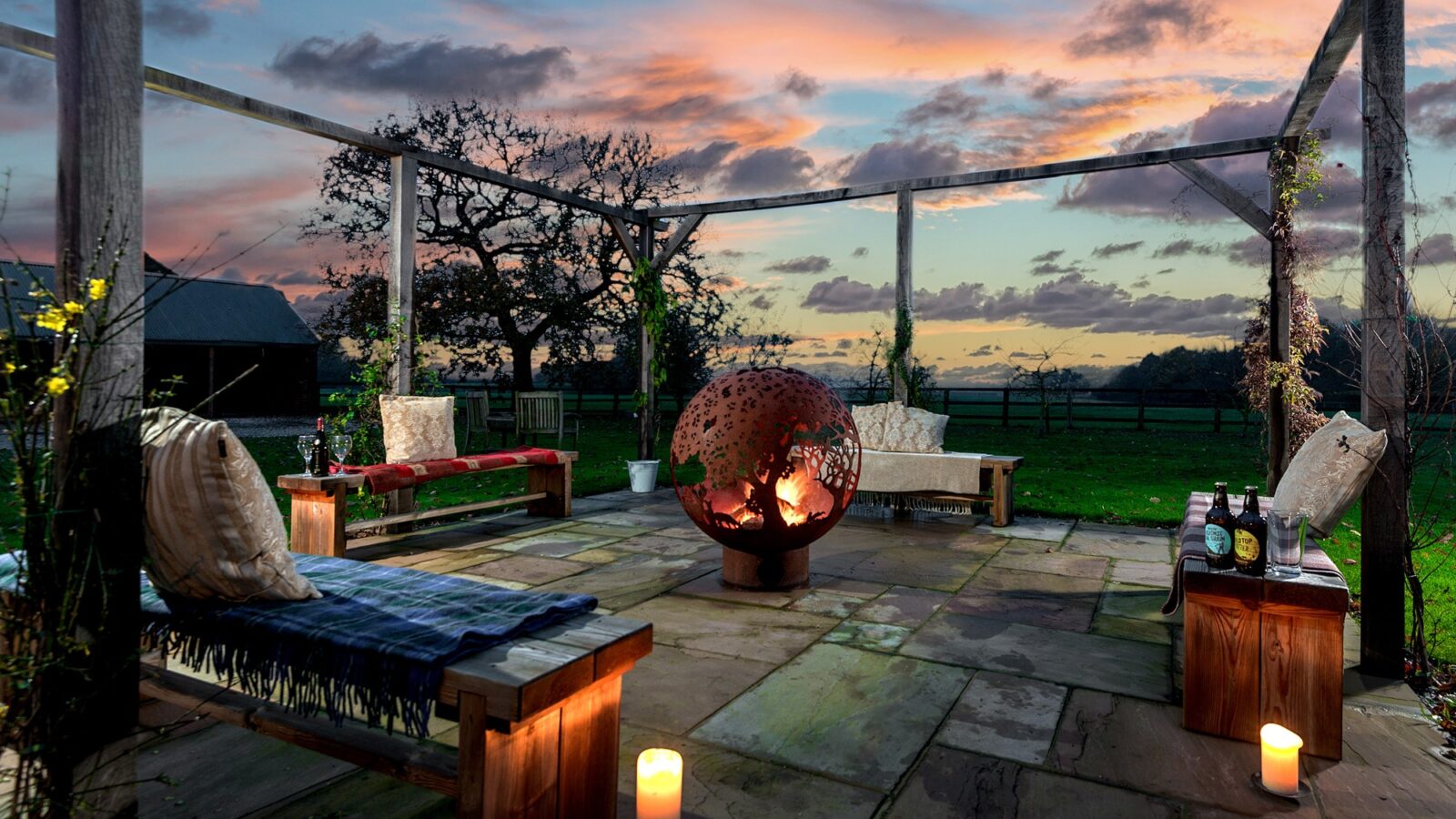 A cozy outdoor patio at sunset at Broadgate Farm Cottages features a central fire pit with a globe design, surrounded by wooden benches with cushions and blankets. Lit candles and drinks on a table add to the inviting ambiance, while trees and open fields are visible in the background.