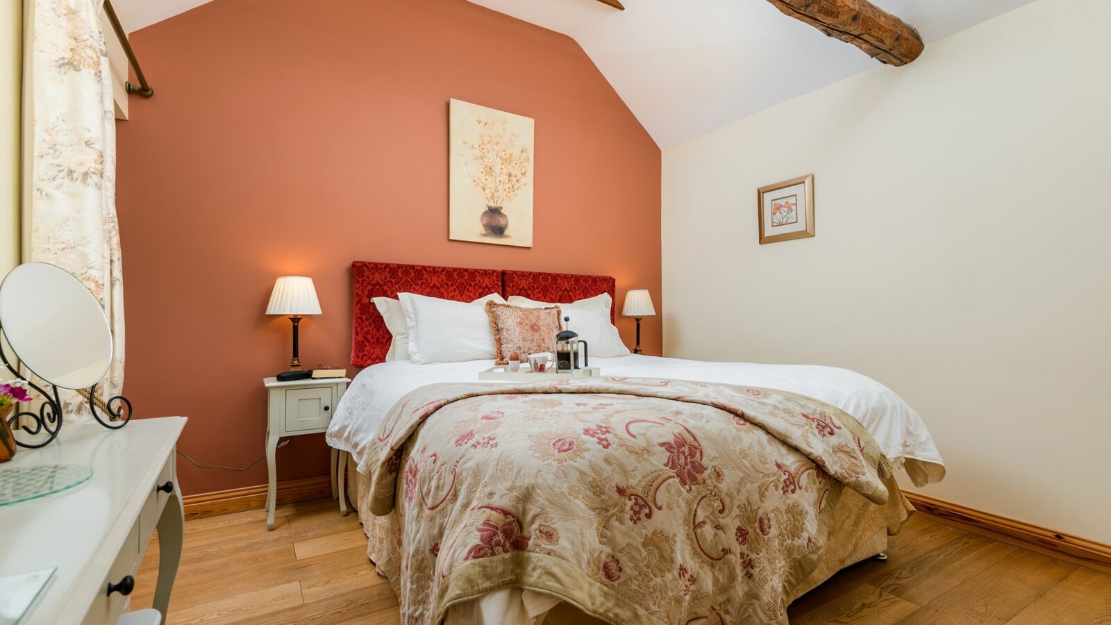 A cozy bedroom at Broadgate Farm Cottages features a double bed adorned with white linens and a patterned throw blanket. The bed has a cushioned red headboard and two bedside tables with lamps. The room boasts wooden flooring, floral artwork, and a slanted ceiling with an exposed wooden beam.