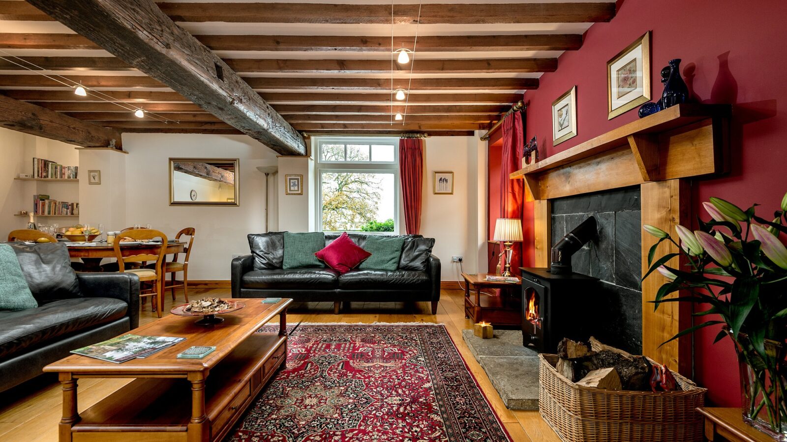 A cozy living room at Broadgate Farm Cottages with exposed wooden beams, a red accent wall, and a black wood-burning fireplace. The room features a dark leather sofa with red cushions, a wooden coffee table, a patterned red rug, and a dining area in the background with bookshelves.