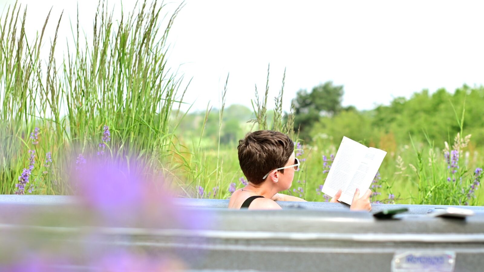 A person with short hair and wearing sunglasses relaxes in the hot tub of the Mercury Eco Cabin, reading a book. Tall grasses and purple flowers surround the hot tub, with a backdrop of greenery and a clear sky.