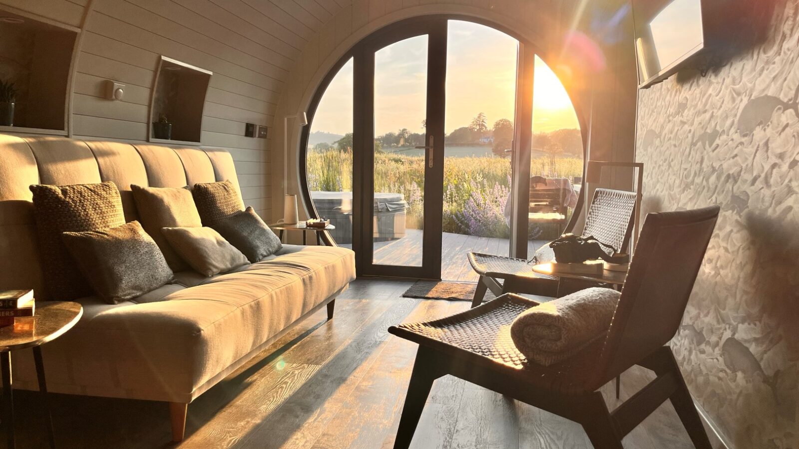 A cozy cabin living area with a curved wooden ceiling features a beige sofa adorned with pillows, two modern chairs, and a coffee table. The room is bathed in warm sunlight streaming through a large arched window with a scenic view of an eco-friendly garden at sunset.