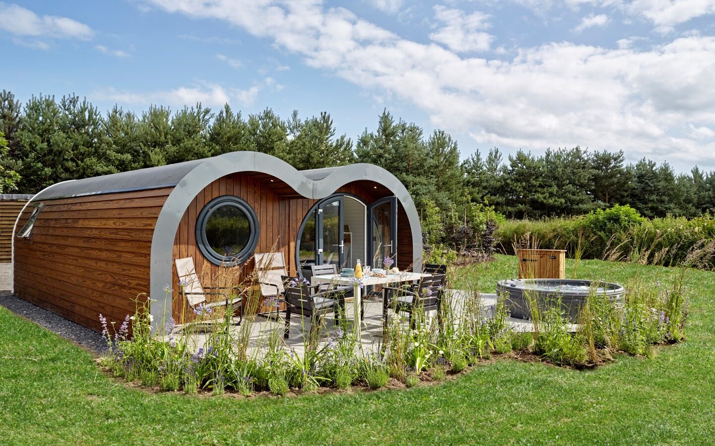 A modern, curved wooden eco cabin with large circular windows sits in a lush green field. The patio features a round dining table with chairs, and a large hot tub. Surrounding the cabin are a variety of plants and trees, with a blue sky and scattered clouds overhead.