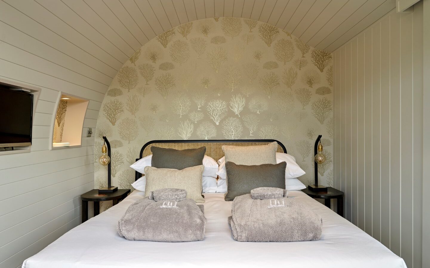 A cozy, cabin-style bedroom with a double bed adorned with white linens and gray throw pillows. Two neatly folded gray towels sit at the foot of the bed. The wall behind the bed features elegant, patterned wallpaper. Two small bedside tables with lamps are on either side.