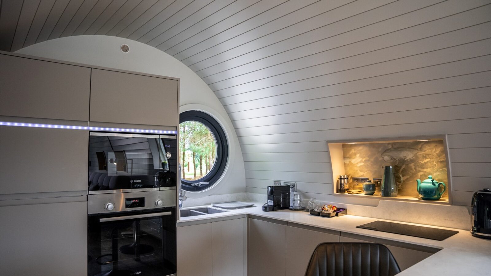 A modern kitchen with curved, white-paneled walls features a round window above the sink, built-in oven and microwave on the left, an eco-friendly coffee machine and toaster in the middle, and shelving with decorative items on the right. A cushioned chair is in the foreground.