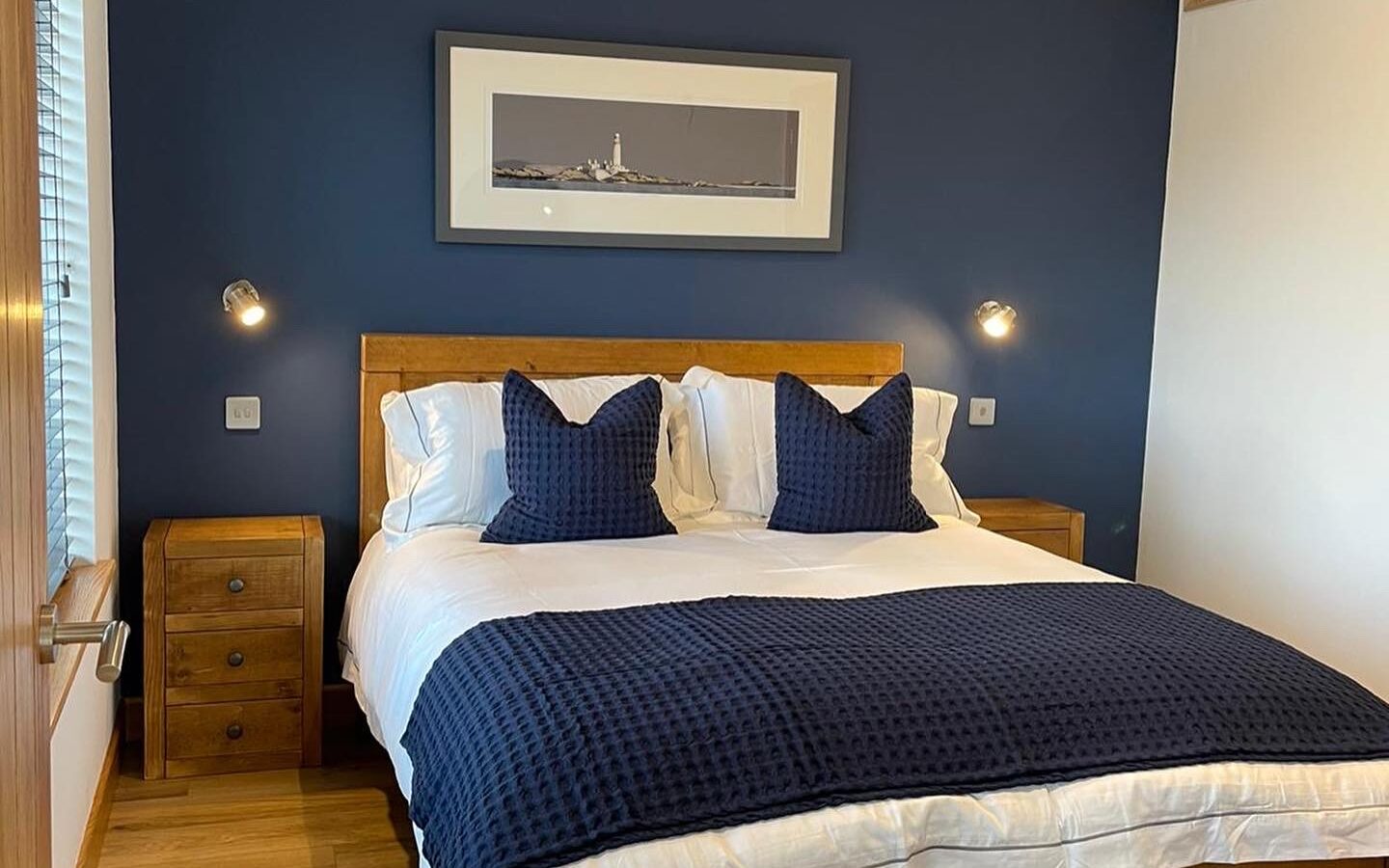 A cozy bedroom reminiscent of lodges, featuring a wooden bed with white bedding and navy blue pillows. A navy blue accent wall behind the bed showcases a framed photograph. Two wooden nightstands with small lamps flank the bed. The room boasts wooden floors and a window on the left.