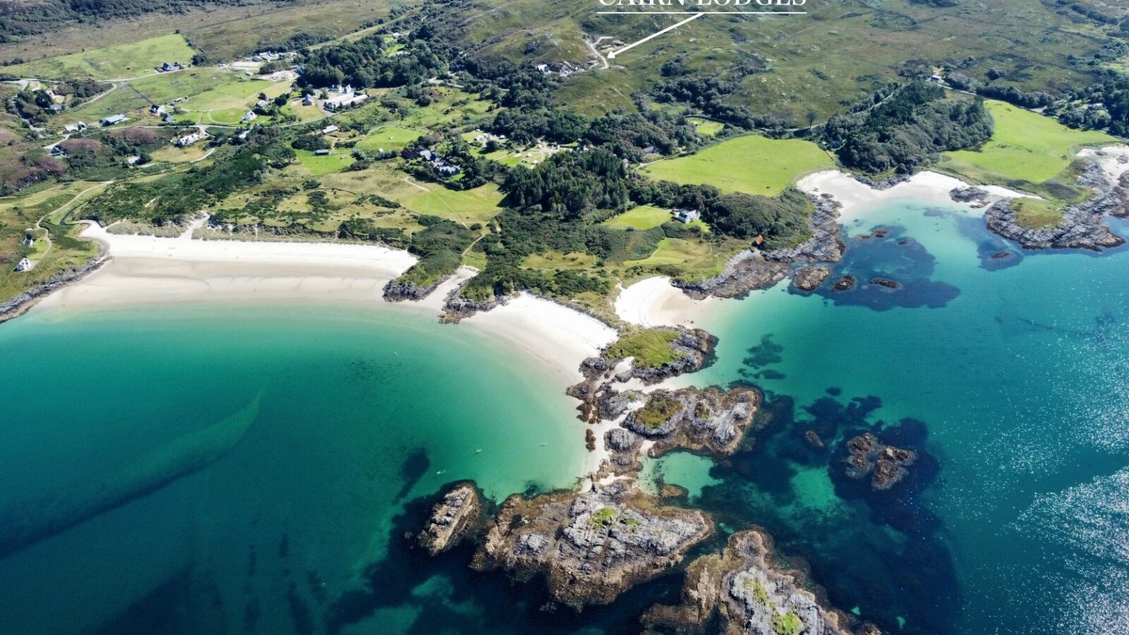 Aerial view of a coastal area with clear turquoise waters, sandy beaches, and rocky outcrops. Green fields and trees surround the coastline. The text 