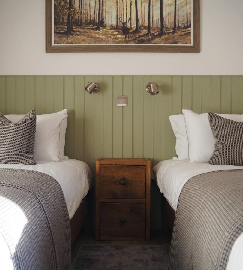 A cozy bedroom in Cairn Lodges features two single beds separated by a wooden nightstand. Each bed boasts white linens, a checkered gray throw, and matching pillows. Above the nightstand, two reading lights are mounted on a green paneled wall with a forest-themed painting above, ideal for Cairn holiday accommodations.