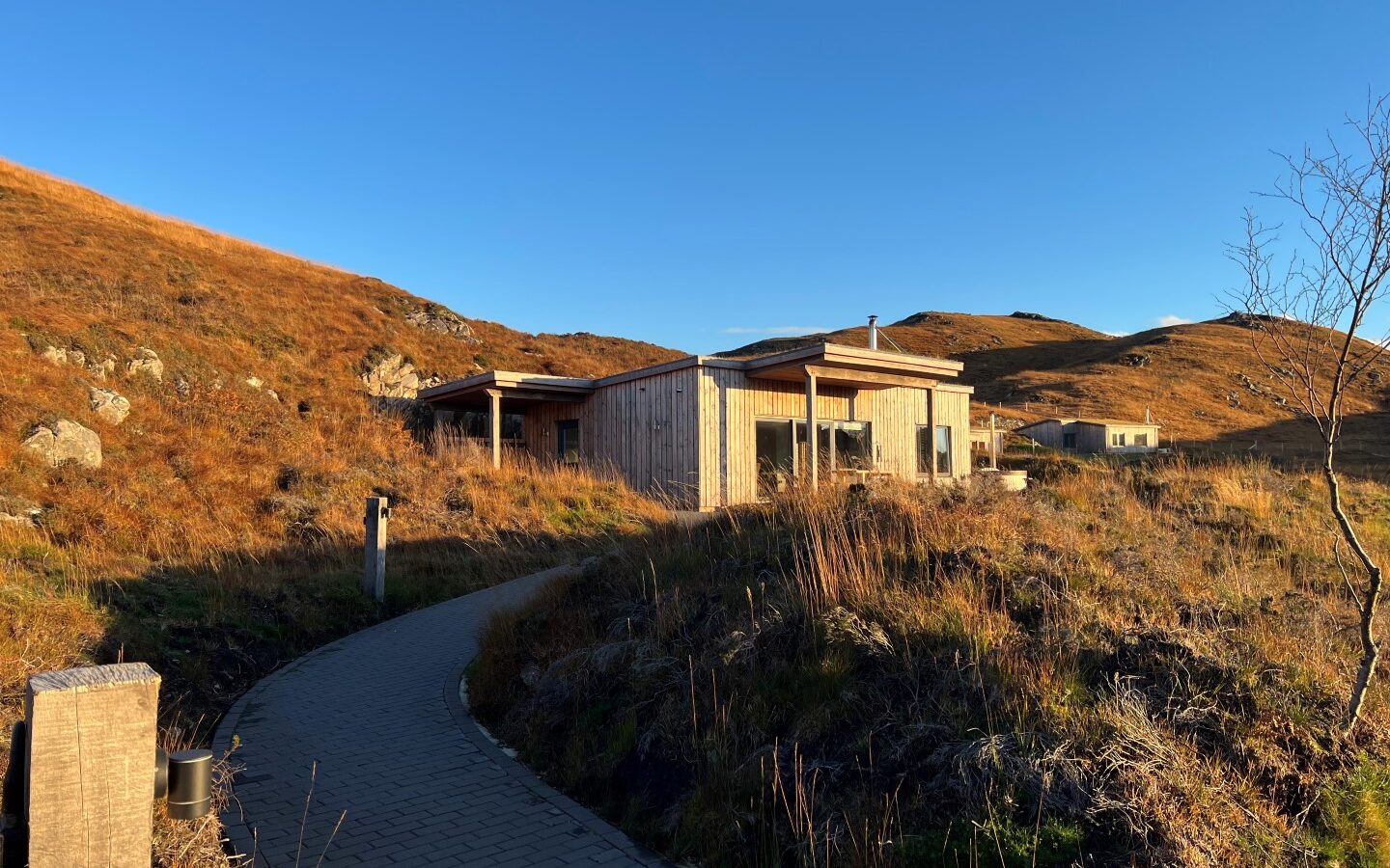 A modern wooden cabin sits on a grassy hillside under a clear blue sky at sunset. A paved pathway leads to the lodge, surrounded by the golden hues of dried grasses and shrubs typical of late autumn. A cairn marks the trail as another cabin is visible in the background.