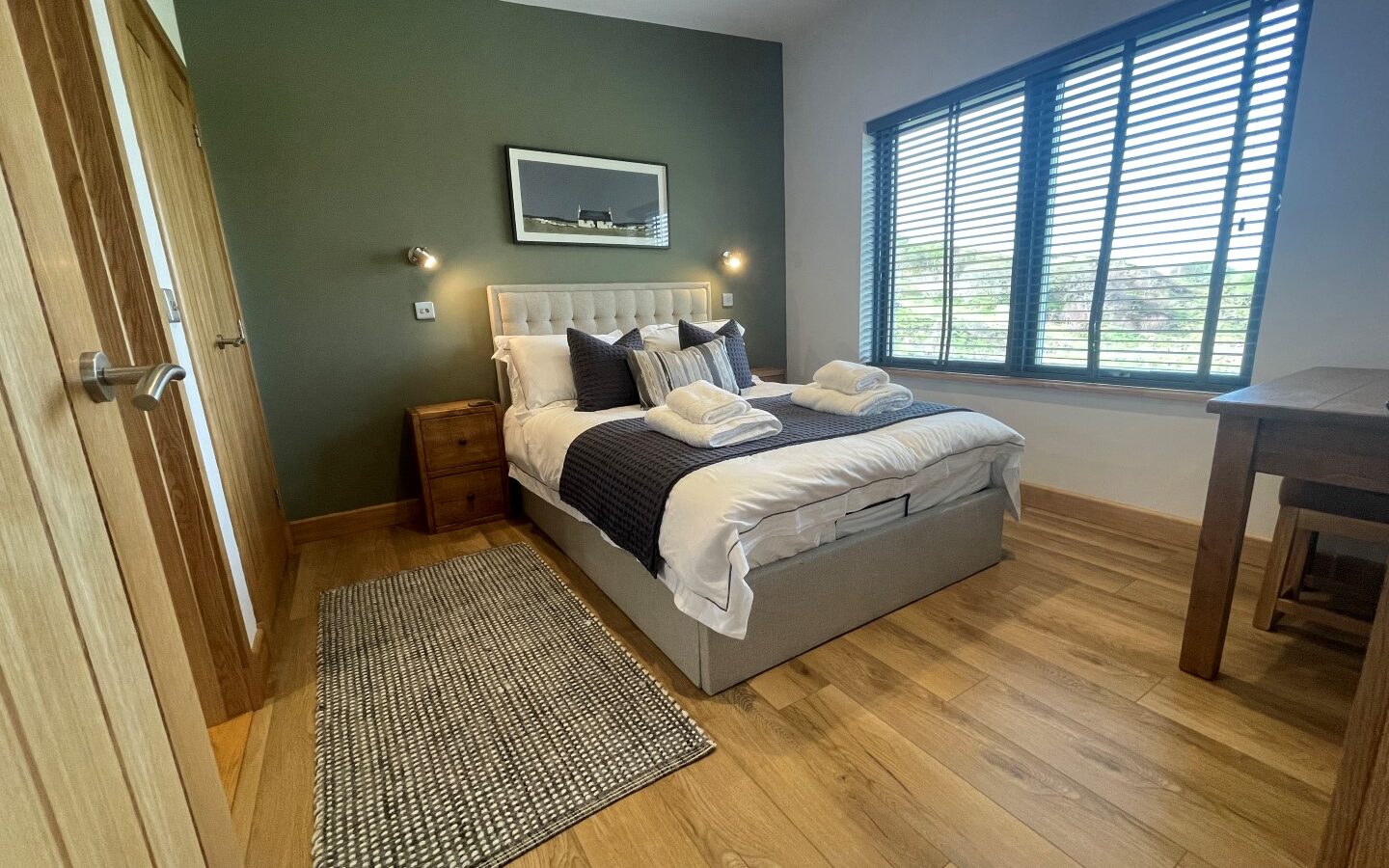 A cozy bedroom in the Cairn Lodges with a double bed featuring white and dark blue bedding, flanked by wooden nightstands with wall-mounted lights. A green accent wall adds color, and large windows with blinds allow natural light. A wooden table and a woven rug complete the room.