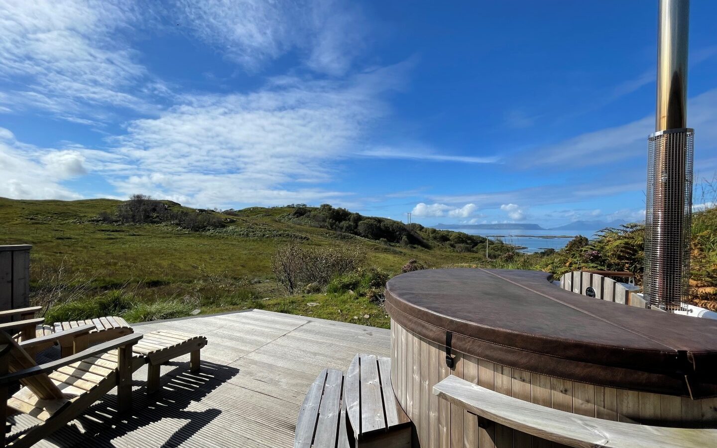 A scenic outdoor view at Cairn Lodges features a wooden deck with seating and a covered hot tub. The deck overlooks a lush green landscape under a partly cloudy blue sky. The horizon offers rolling hills and a glimpse of the ocean in the distance.