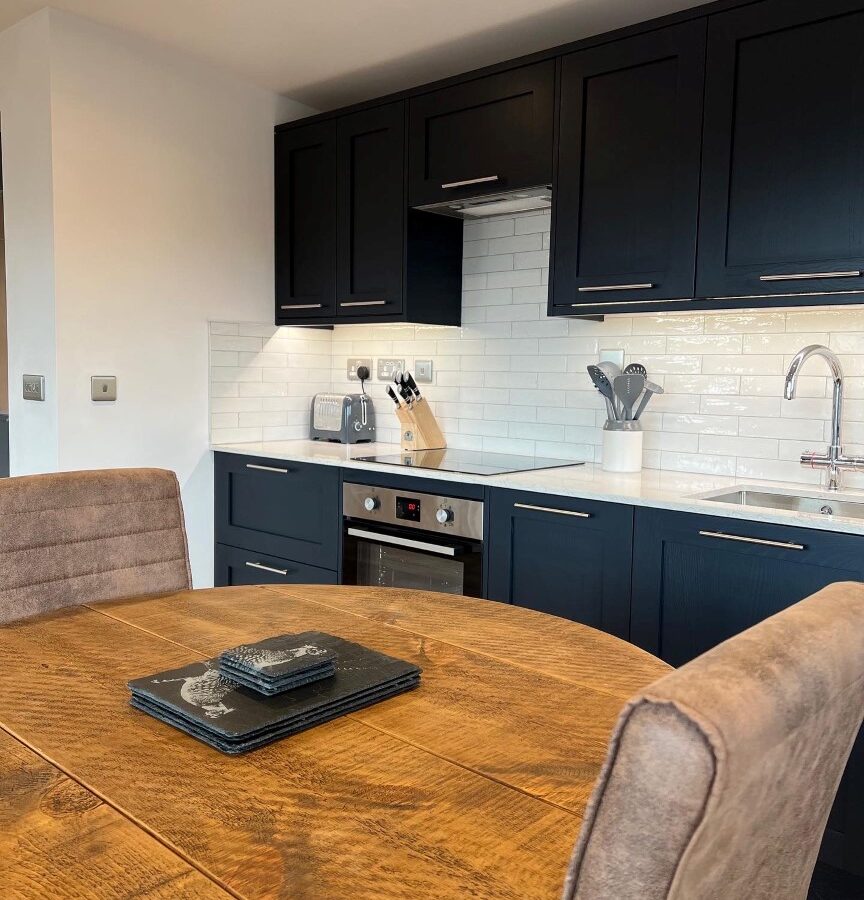 A modern kitchen at Cairn Lodges features black cabinets, a white tiled backsplash, and stainless steel appliances including an oven and toaster. A wooden dining table with leather chairs sits in the foreground, while a knife block and utensil holder adorn the countertop.