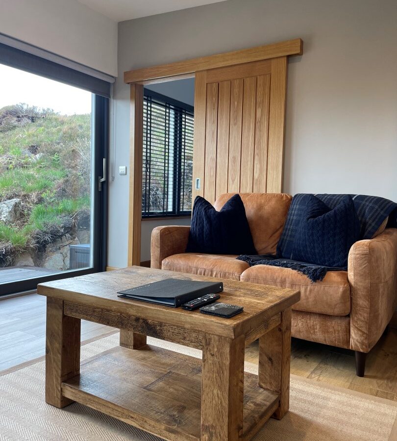 A cozy living room at Cairn Lodges features a brown leather sofa with navy pillows against a light gray wall. The rustic wooden coffee table holds a closed laptop and remote controls. A large window reveals a green, rocky landscape outside, and a wooden sliding door is partially closed.