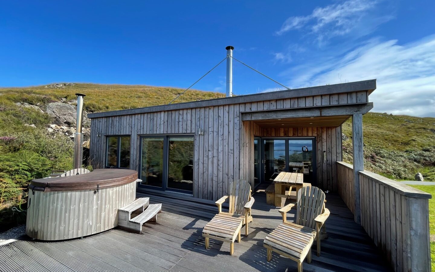 A modern cabin with a wooden exterior features large glass doors, a hot tub, and several Adirondack chairs on the deck. Nestled among lodges on a grassy hillside under a bright blue sky with some wispy clouds, it offers a scenic view of the ocean in the distance.