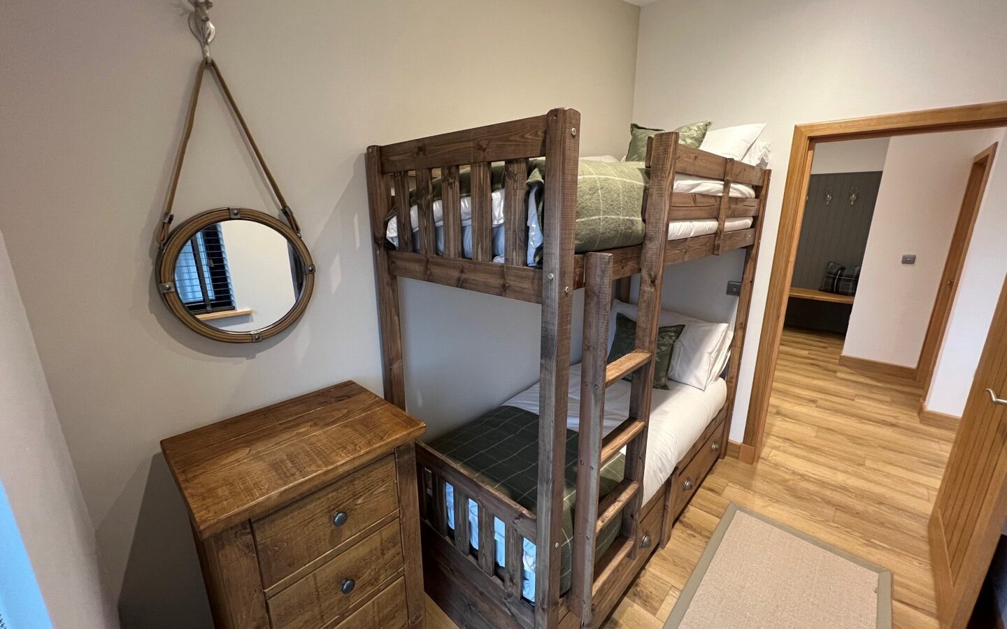 A cozy bedroom in Cairn Lodges features a wooden bunk bed with checkered bedding, a matching chest of drawers, and a round mirror mounted above. The room has wooden flooring and beige walls, with a doorway leading to another room.