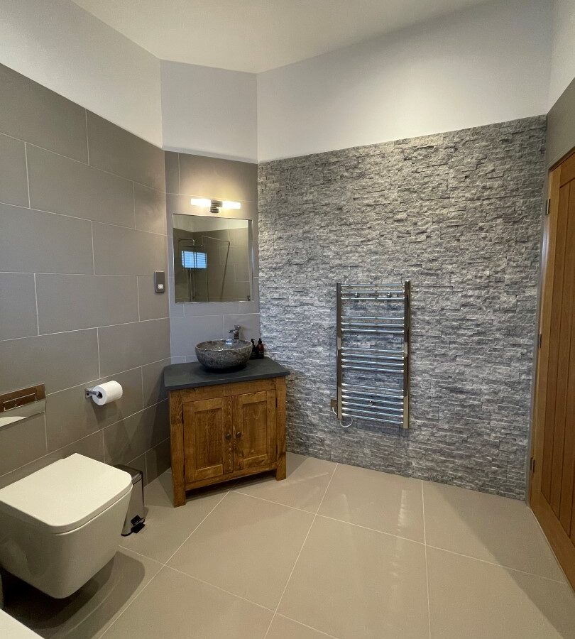 A modern bathroom at Cairn Lodges with grey and beige tones features a wall-mounted toilet, a wooden vanity with a vessel sink and mirror above it, and a heated towel rail on a textured stone accent wall. The floor is tiled, and the lighting is recessed in the ceiling.